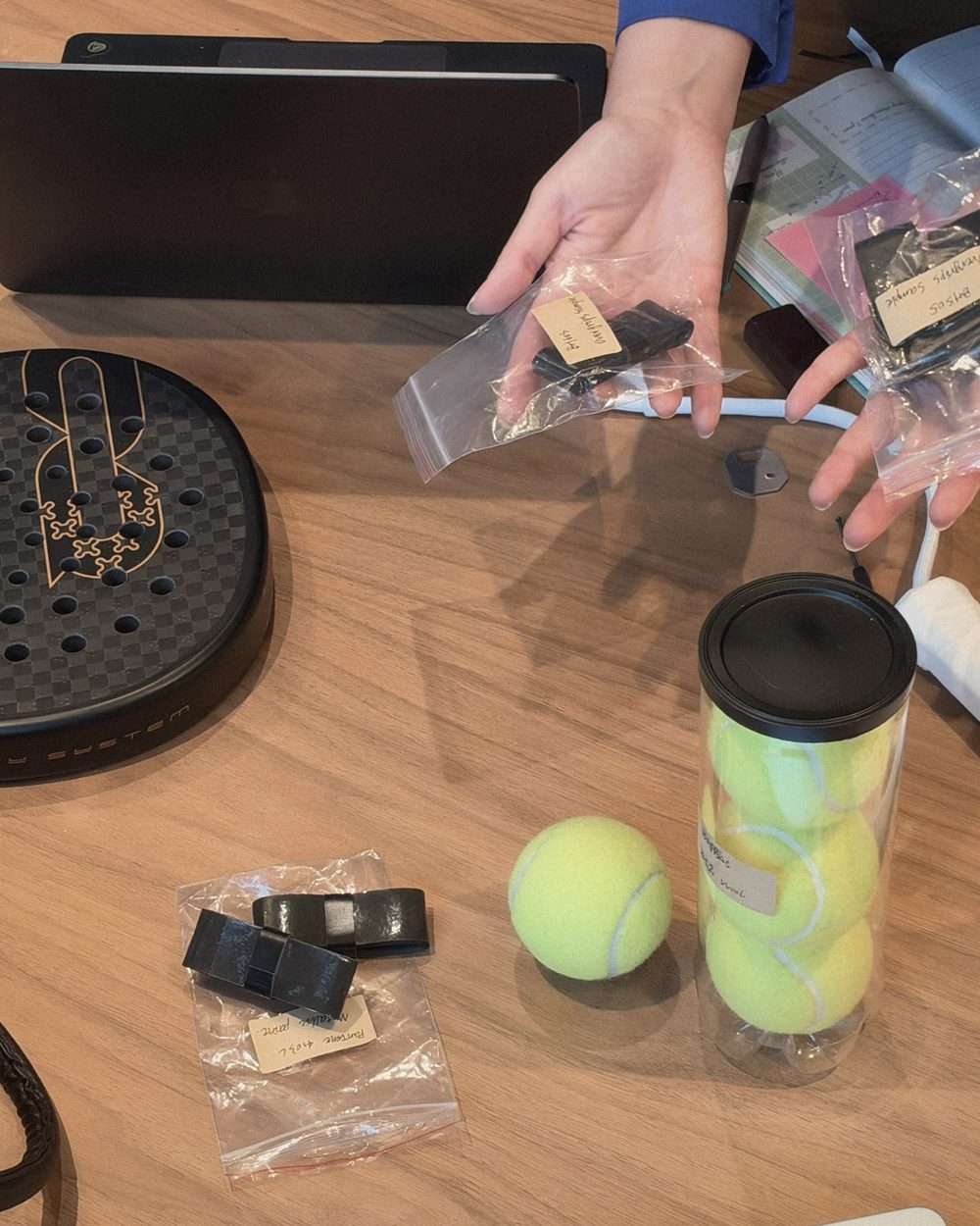 Hands holding labelled overgrip samples over APEX, with balls and grips on the table