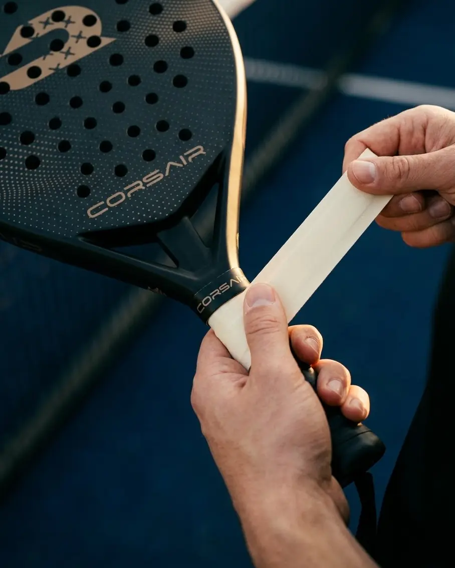 Two hands winding a cream Corsair grip onto APEX at the throat, on a blue court in low evening light
